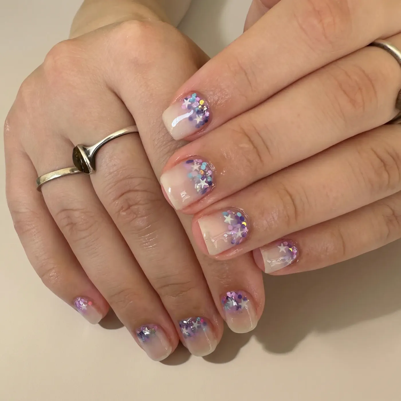 Melkehvit nude base med lilla, blå og rosa glitter og sølvstjerner i myk overgang mot tuppen
