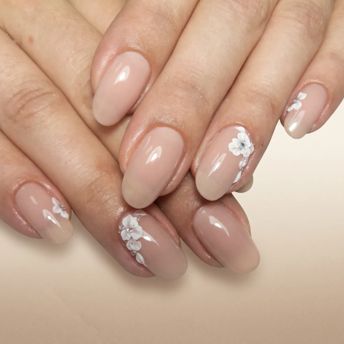 Ovale nude beige negler med hvite 3D-blomster i gel langs neglekanten og små sølvstrass